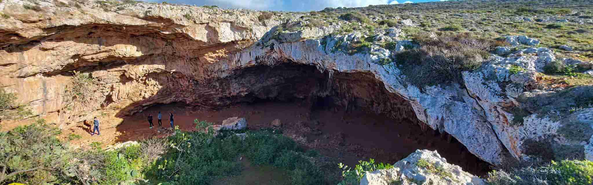 Wide limestone cave in Malta with a large arched entrance and several visitors standing inside, highlighting the scale of the natural rock formation