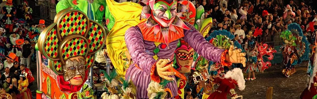Large, colourful Carnival float with masked jester figures and performers parading through a crowded street during Malta Carnival celebrations at night