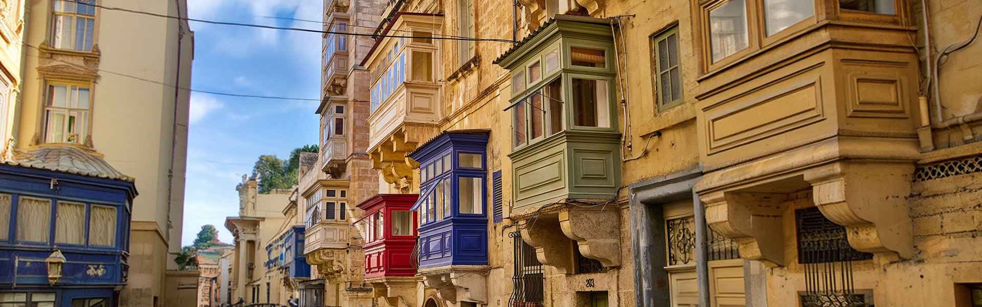 Colourful traditional Maltese gallariji in red, blue and green line the upper floors of honey-coloured limestone buildings along a narrow Valletta street.