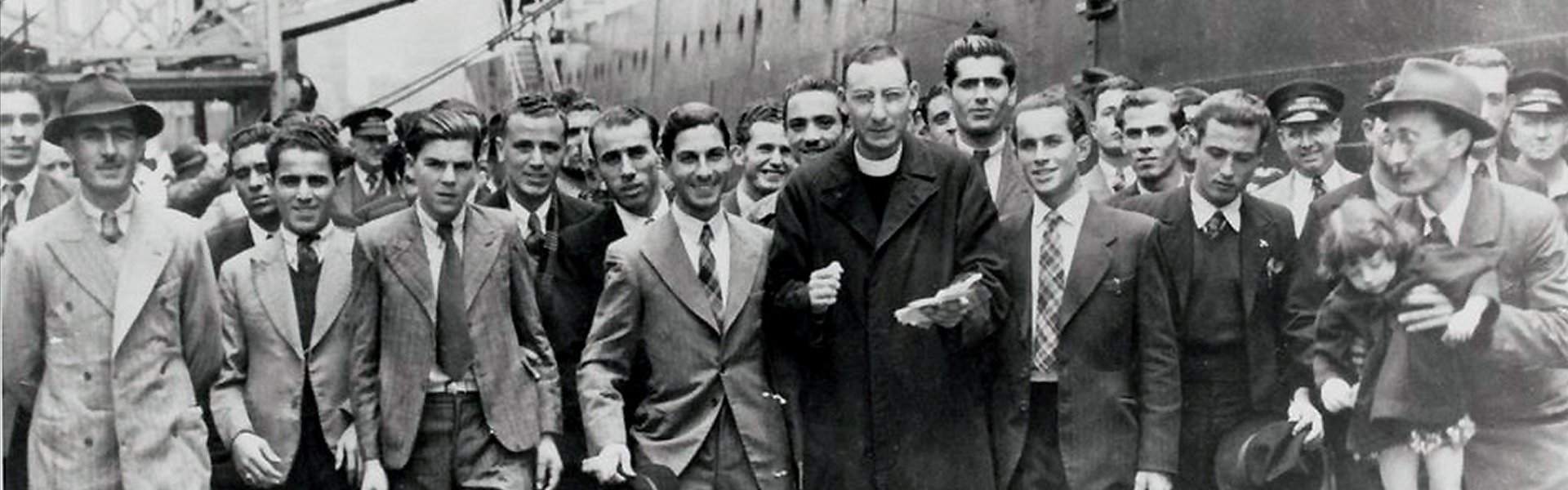 Black and white historical photograph of a large group of Maltese men walking together near a docked ship, accompanied by a Catholic priest in clerical dress, likely at a port during the mid-20th century emigration era.