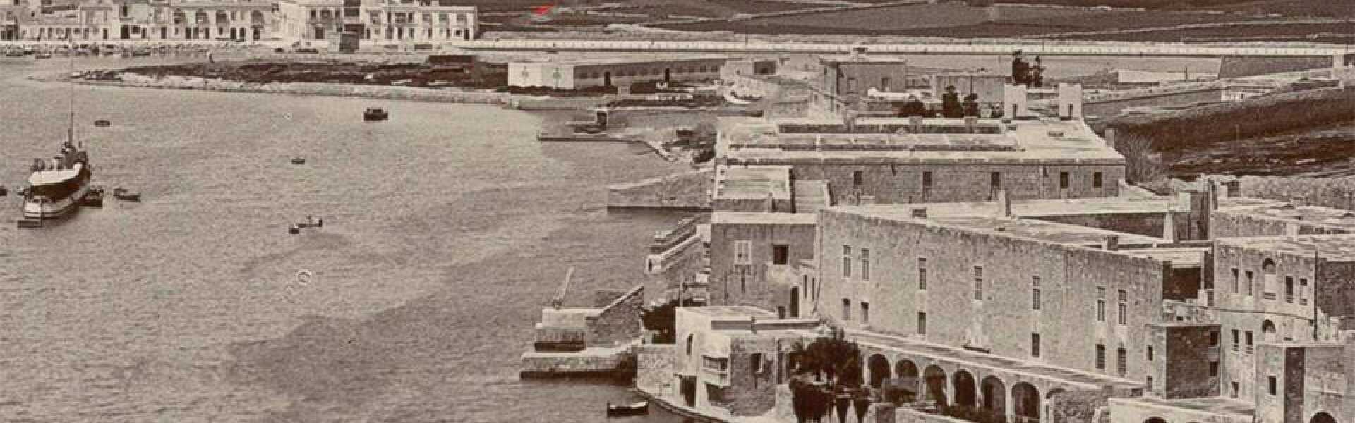 Wide sepia-toned panoramic photograph of Marsamxett Harbour in Malta, showing the historic Lazzaretto quarantine complex on Manoel Island with its long stone arcades and waterfront buildings. Small boats sit moored in the calm harbour, with terraced field