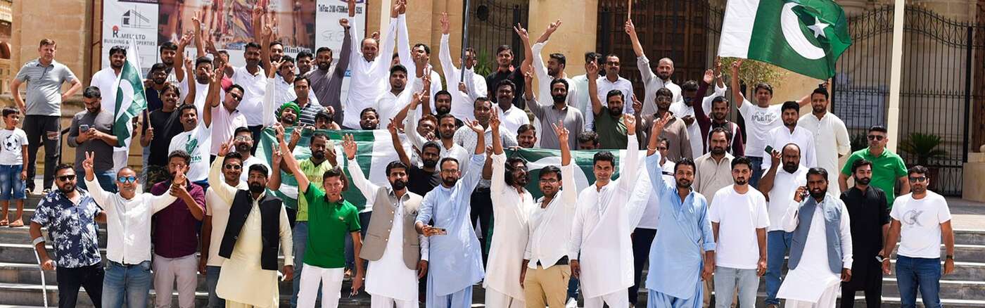 A large group of Pakistani men gathered on the steps of a Maltese limestone building, celebrating with raised arms, peace signs, and Pakistani flags  the distinctive green and white national flag visible prominently on both sides of the crowd. Many are d
