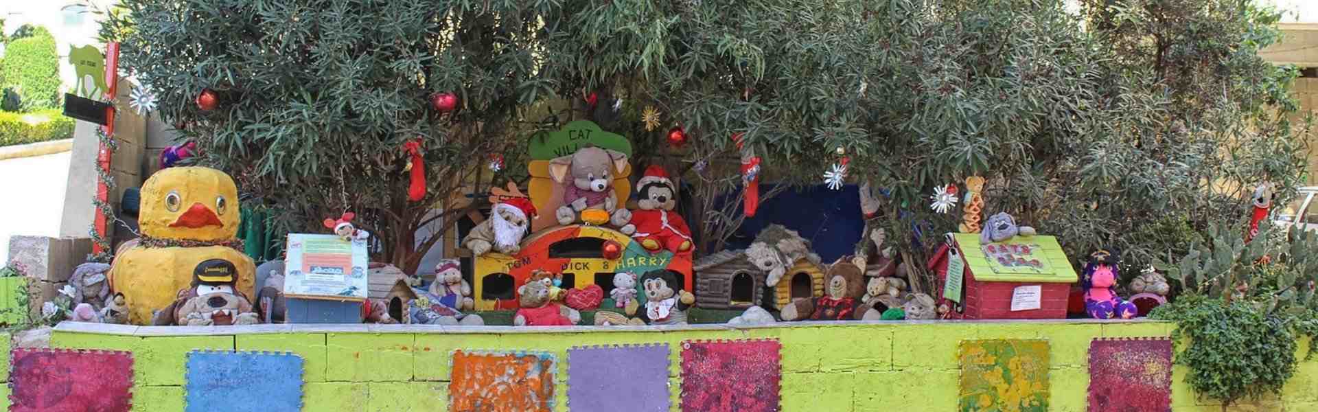A colourful outdoor Cat Village display decorated for Christmas, featuring stuffed animal toys, miniature wooden houses, a yellow duck sculpture, and a 