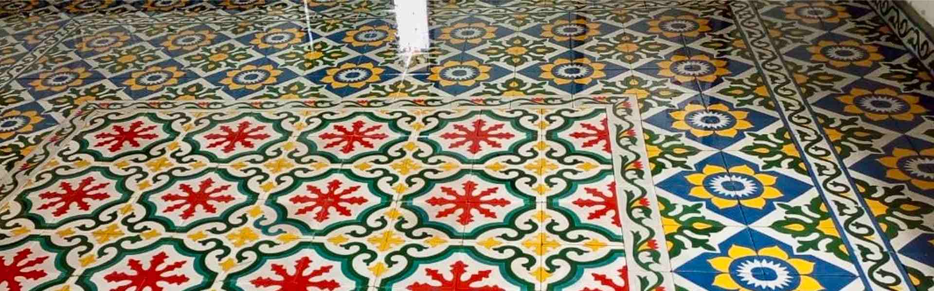 Wide banner photo of a glossy Maltese cement tile floor showing a red star-burst pattern with green scrollwork bordered by a vine trim, set beside a blue diamond pattern with yellow sunflower rosettes and green leaf motifs.