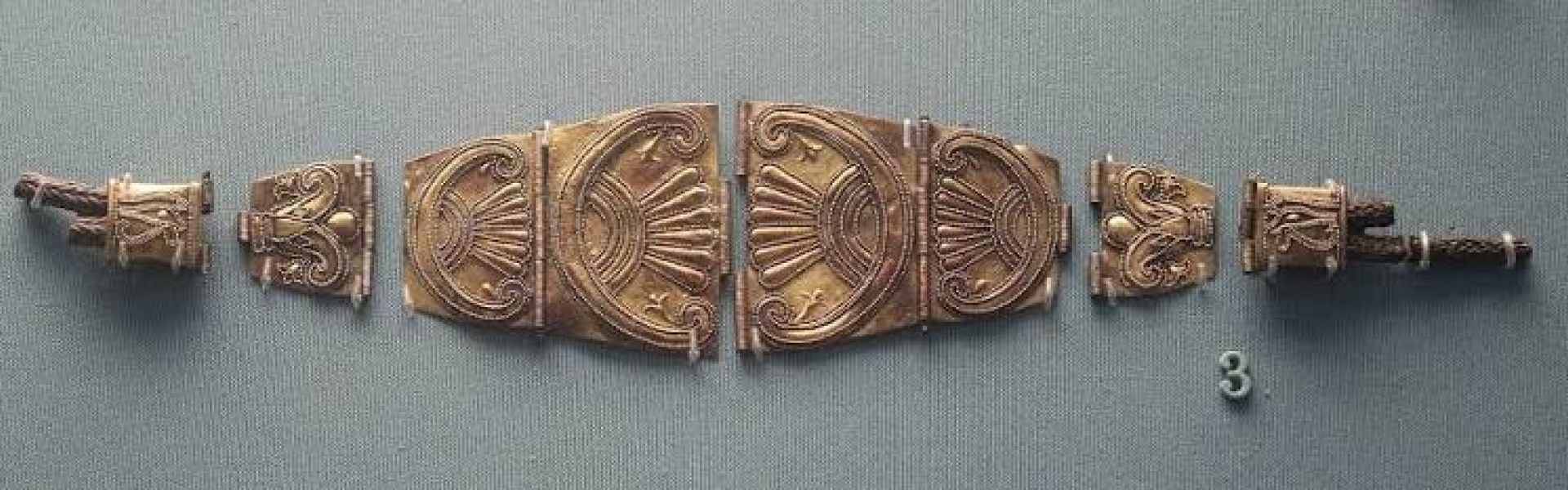 Ornate bronze and gold jewellery fragments from the Phoenician era, decorated with scroll and fan-shaped palmette motifs, displayed on a grey museum backdrop with an item number tag.