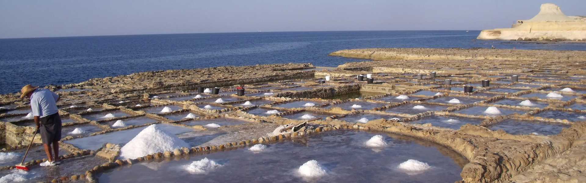 Wide banner of the Xwejni salt pans in Gozo, showing a worker in a straw hat raking harvested sea salt into piles across the rock-cut grid, with the Qolla l-Bajda headland visible on the horizon.