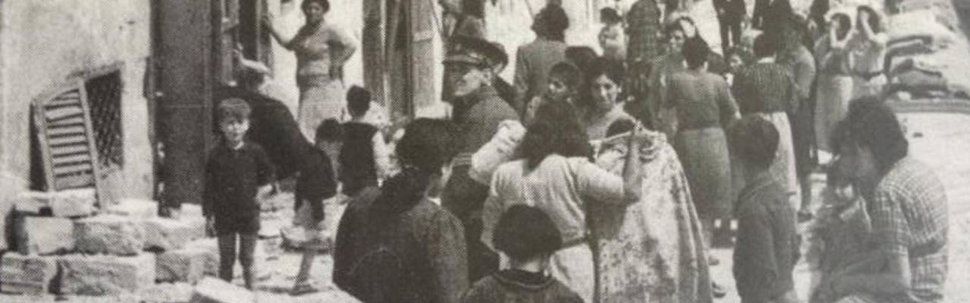 Close-up of a crowd on a rubble-lined Maltese street, with a uniformed officer, women, and children gathered outside damaged whitewashed buildings and stacked masonry blocks, consistent with WWII-era Malta.