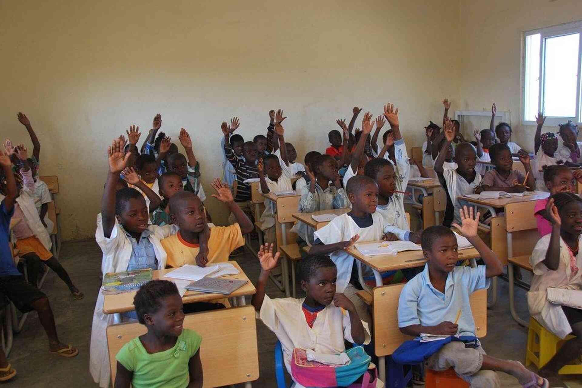A crowded classroom of primary school students eagerly raise their hands at their wooden desks, with open notebooks and textbooks in front of them, in a school in sub-Saharan Africa.