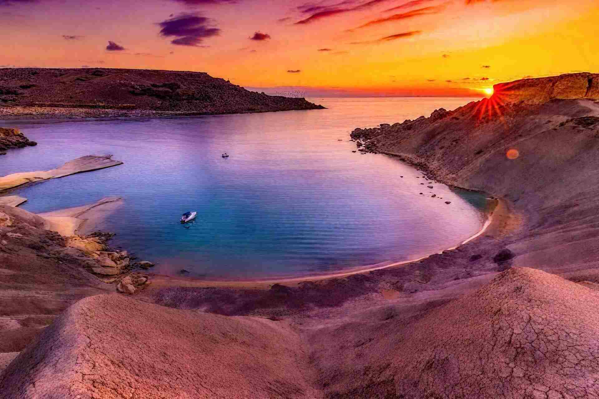Aerial view of a secluded bay in Malta at sunset, with vivid purple and orange skies reflecting over turquoise water and cracked limestone foreground