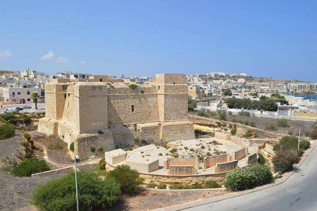 Aerial view of a historic Maltese limestone fort with thick defensive walls and a semi-circular battery, surrounded by scrubland with a coastal town visible in the background.