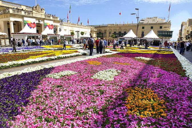 Vibrant floral carpet of pink, purple, white, yellow and orange flowers filling St George's Square in Valletta during the Malta International Flower Festival, with crowds browsing market stalls and historic Baroque buildings in the background