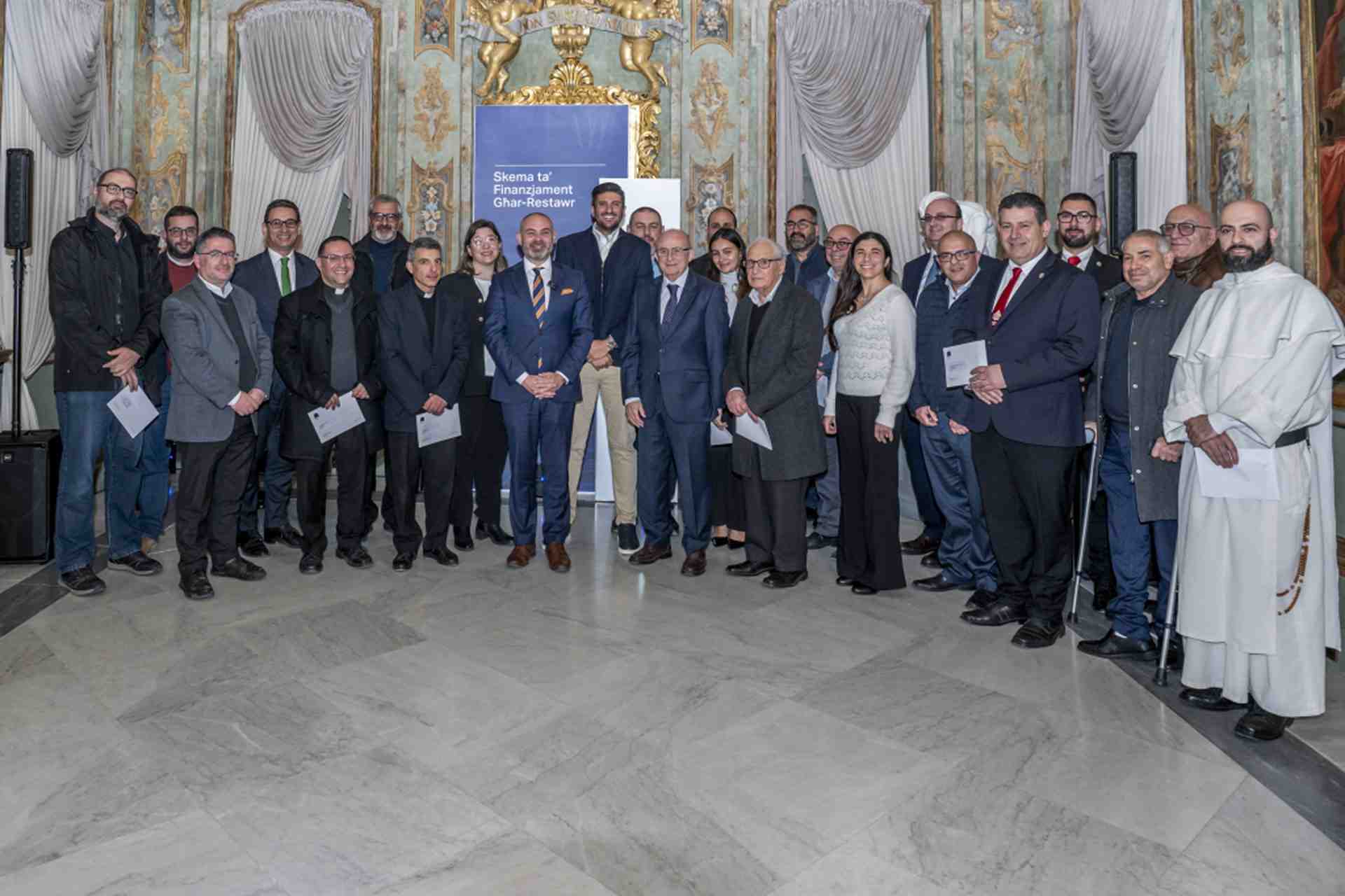 A large group of people posing for a photo indoors in an ornate hall. A blue banner behind them reads: Skema ta' Finanzjament Għar-Restawr