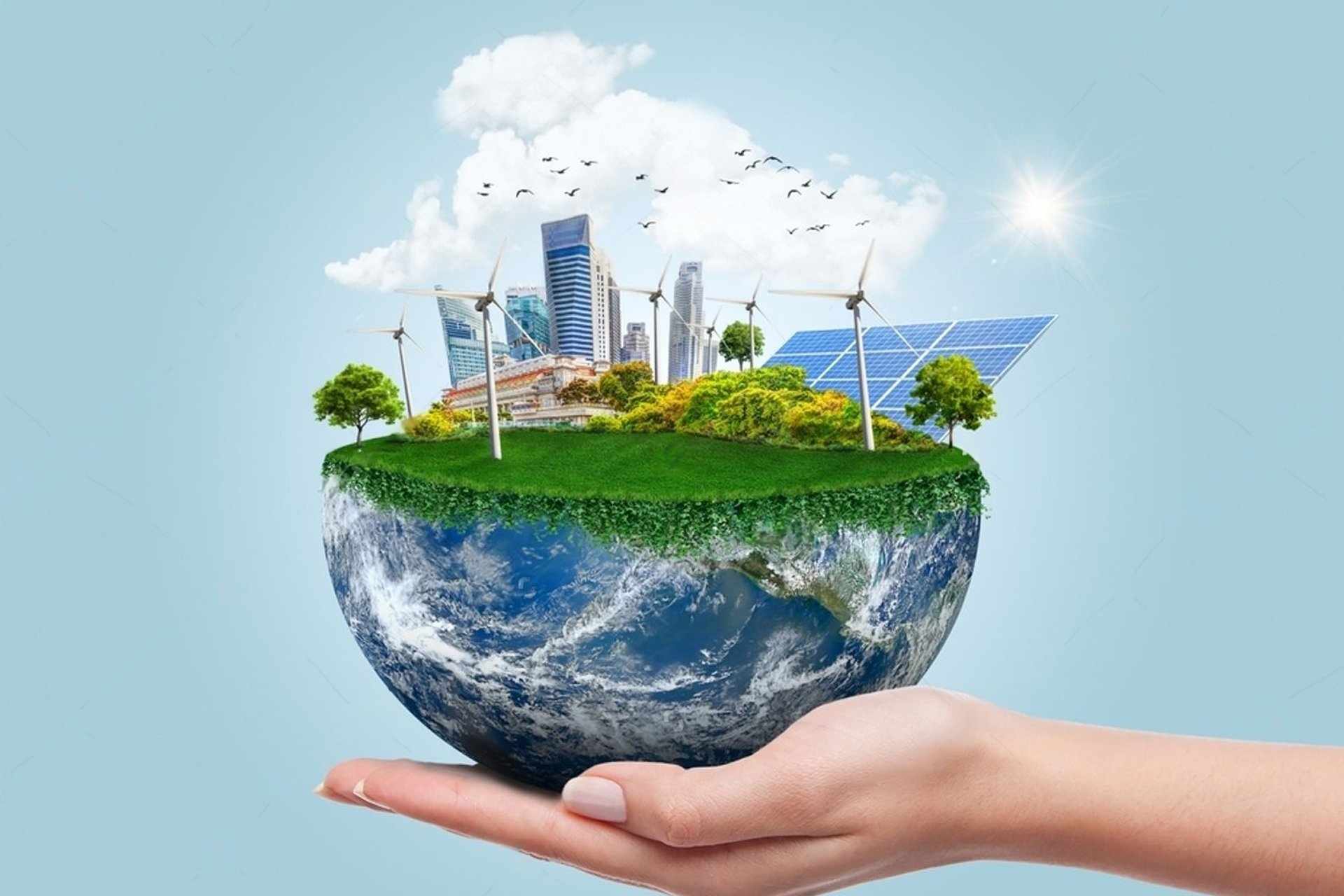 A hand holding a half-globe topped with a green cityscape, wind turbines, solar panels, and trees under a sunny blue sky, representing sustainable development.