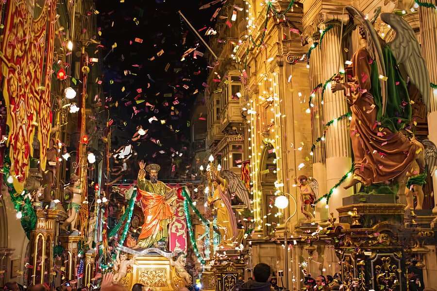 A Maltese village feast procession at night with a gilded saint's statue carried through a confetti-filled street decorated with lights, flags, and ornate religious figures.