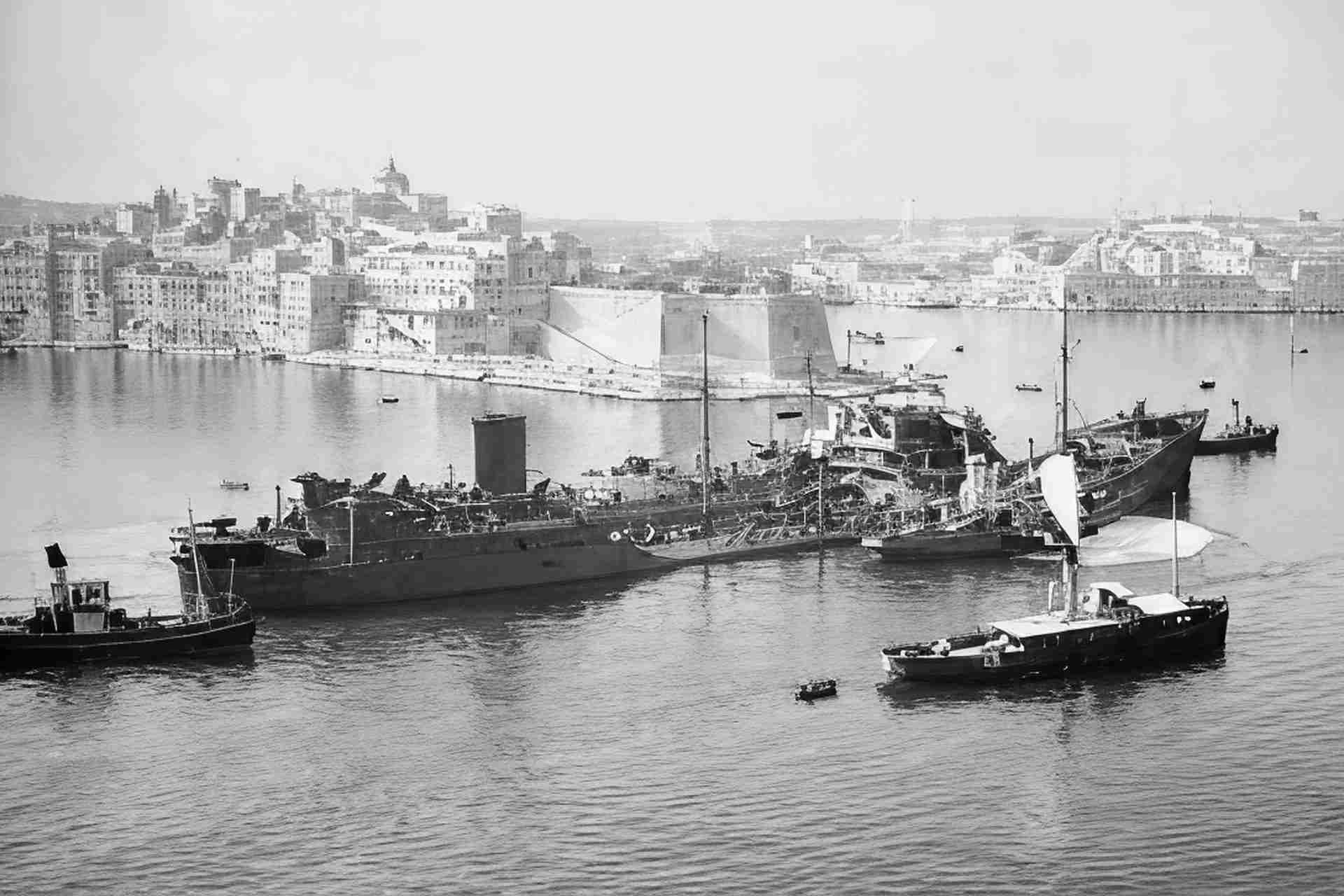 Black and white photograph of a badly damaged merchant vessel being escorted into the Grand Harbour, Valletta, Malta, with the Three Cities visible in the background
