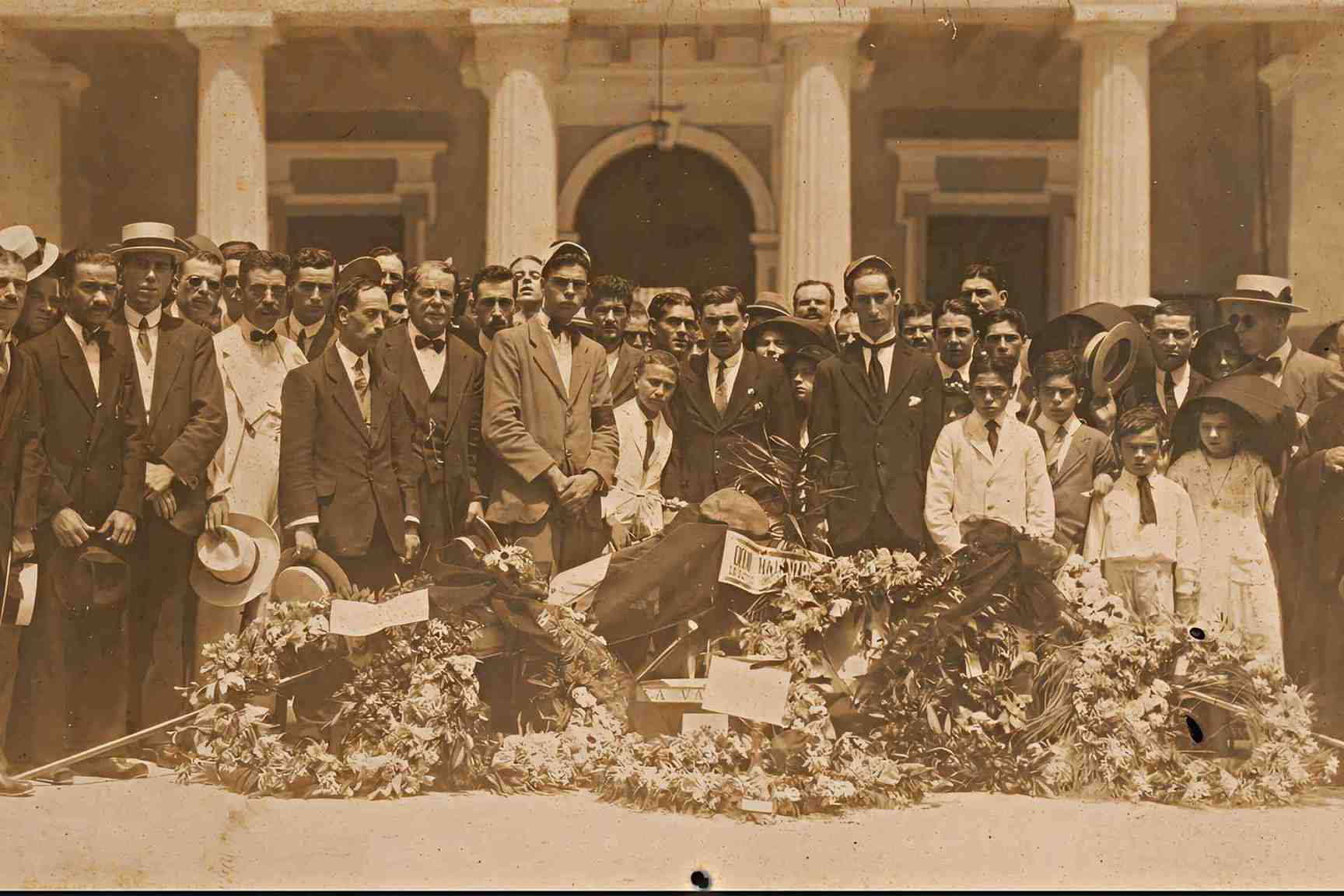 Sepia photograph of a large crowd gathered in front of a colonnaded building in Malta, with men in early 20th-century suits holding hats and floral tributes
