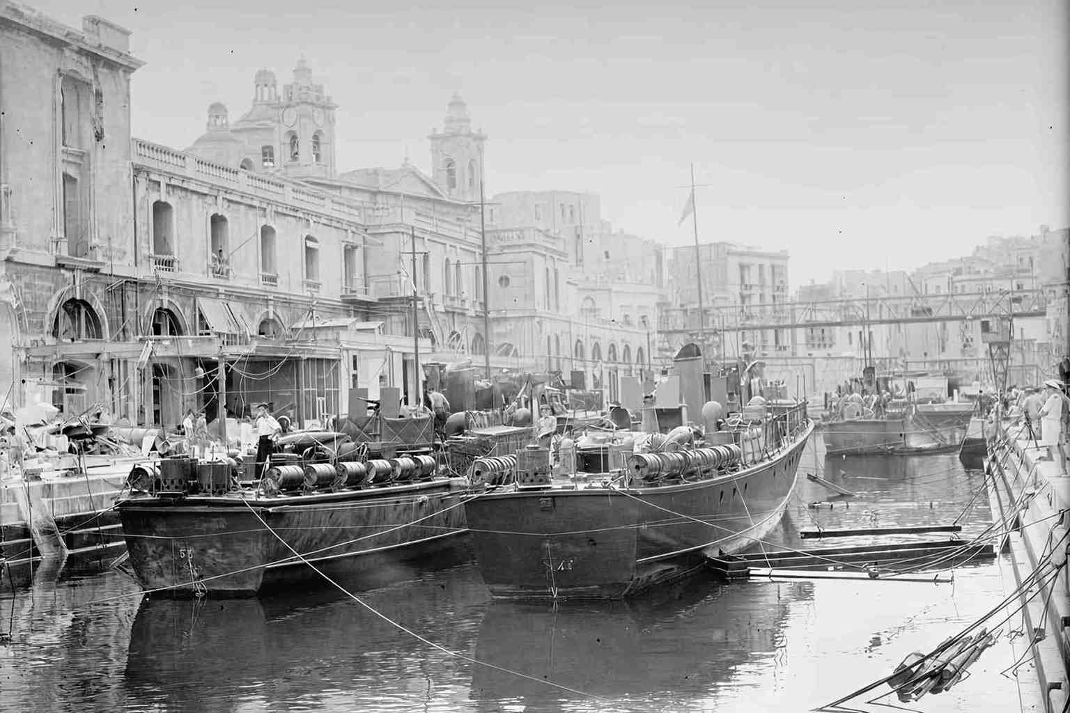 Historic black-and-white photograph of torpedo boats moored in Dockyard Creek, Malta, with Vittoriosa waterfront buildings and church domes visible in the background