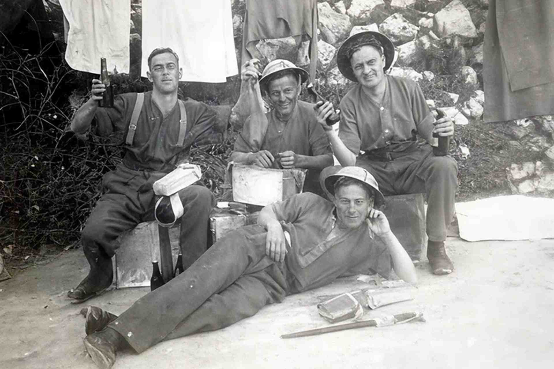 Four WWII soldiers relaxing outdoors holding beer bottles, with laundry hanging on a line behind them and military equipment on the ground