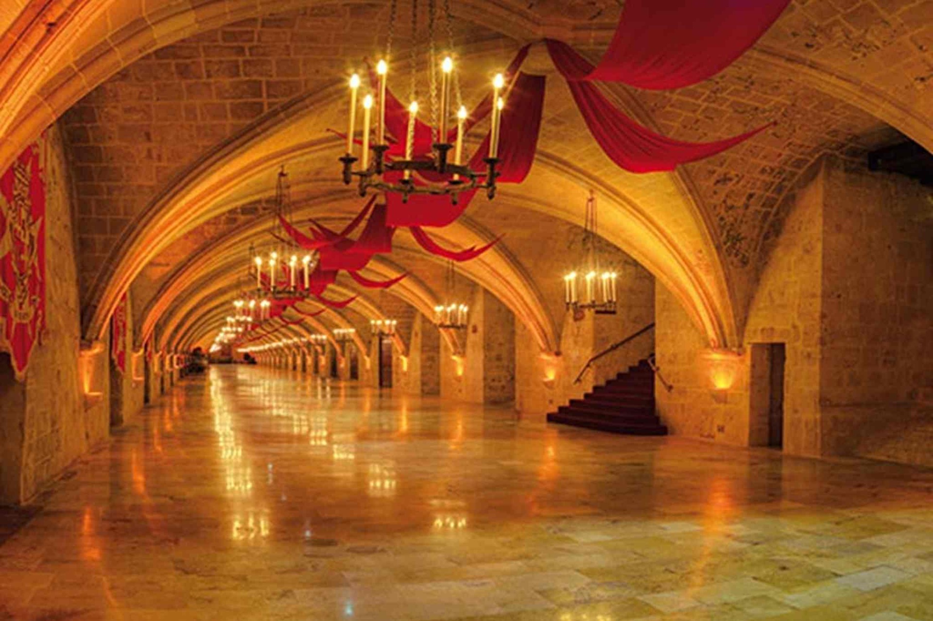 Long vaulted hall with chandeliers, red banners and warm lighting inside a historic palace armoury.