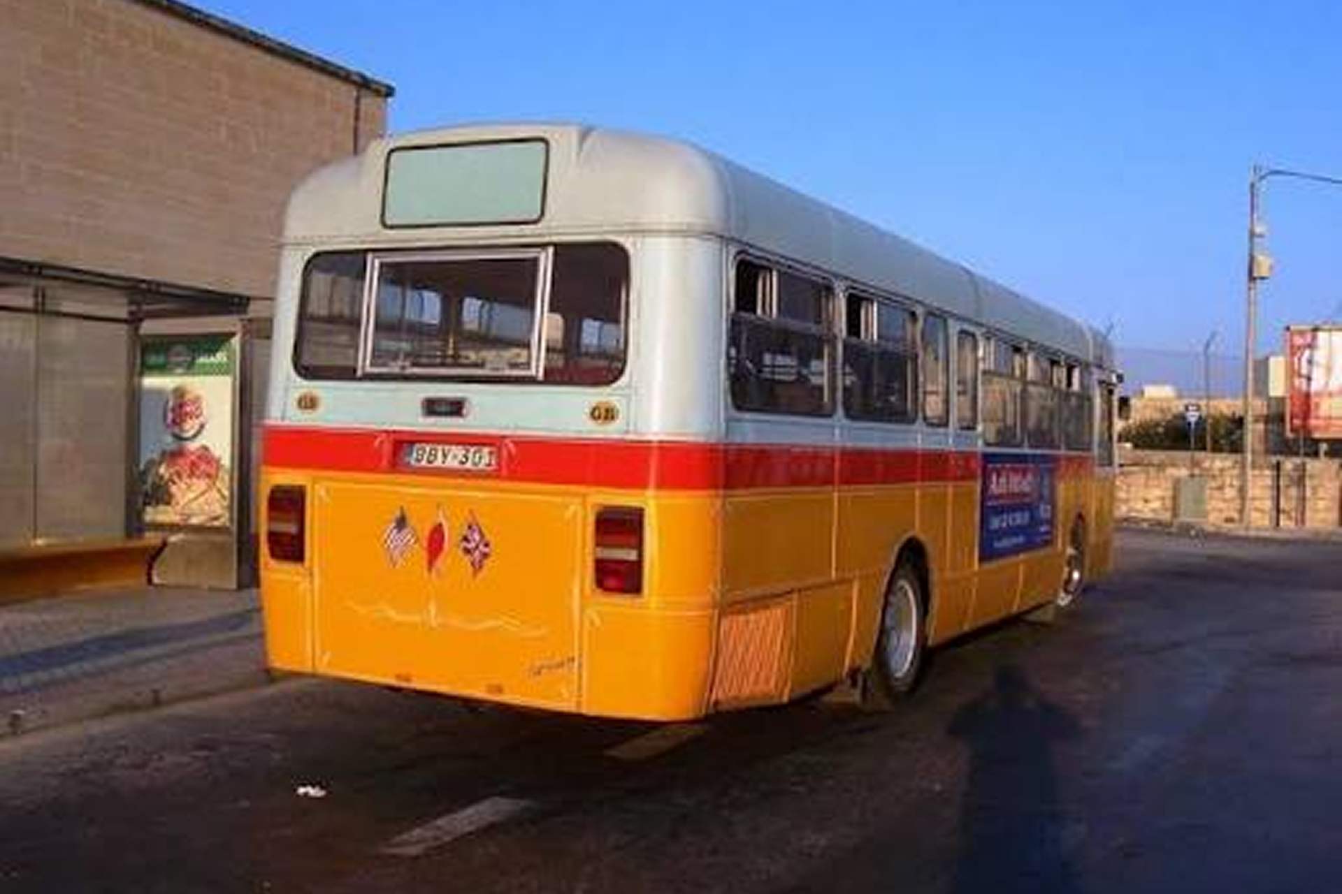 Rear view of a yellow, white and red Maltese bus with small national flag decals, parked near a bus shelter with an