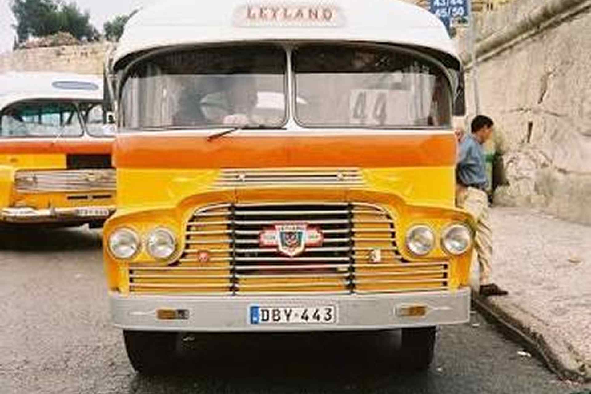 Front view of an orange and yellow vintage Leyland bus displaying route number 44, with another Maltese bus and a man standing beside it in the background.