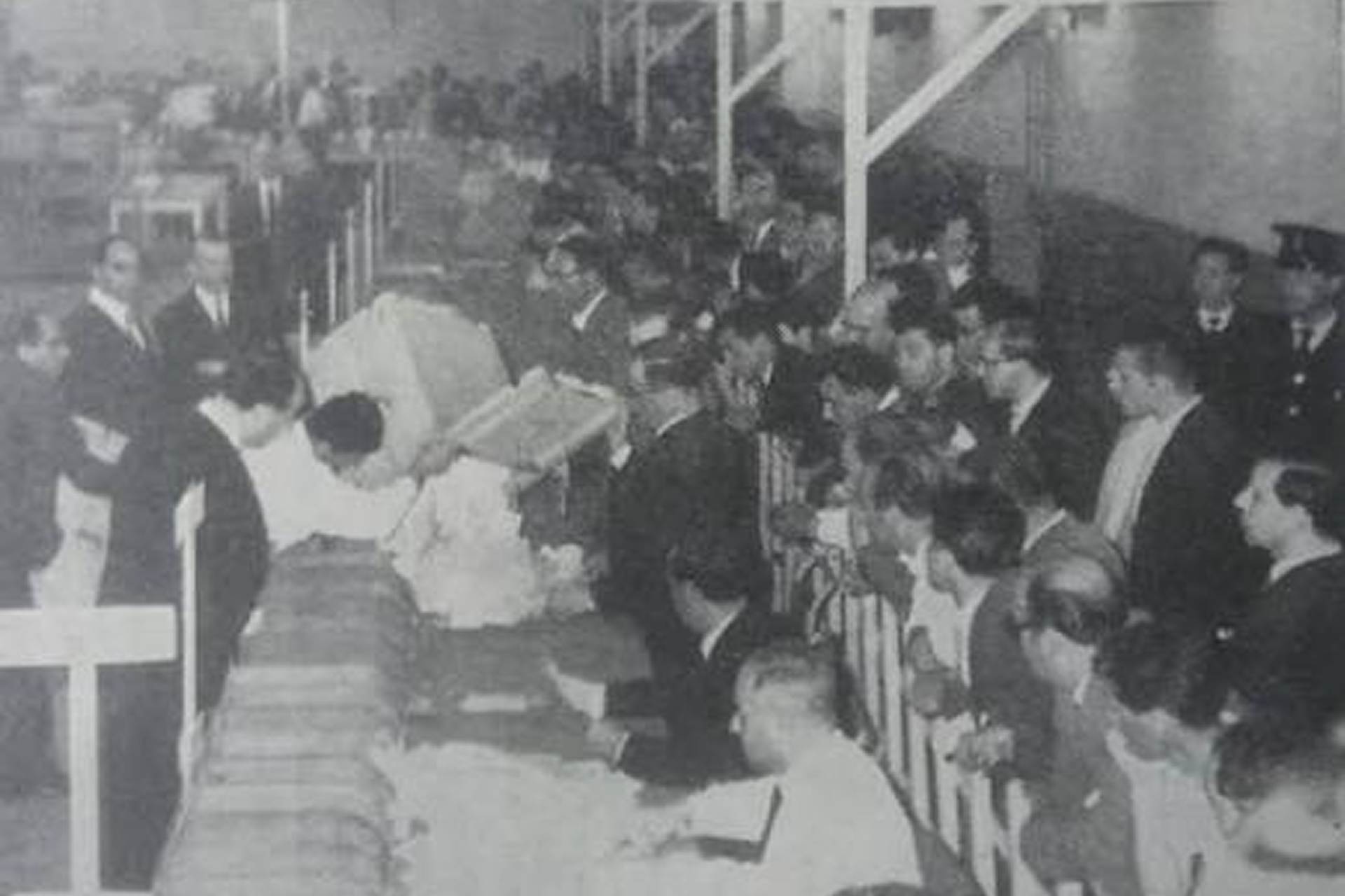 Black and white photograph of the 1956 Malta integration referendum count, with ballot boxes emptied onto a long table as officials sort papers and rows of scrutineers, police officers and onlookers watch from behind a barrier.