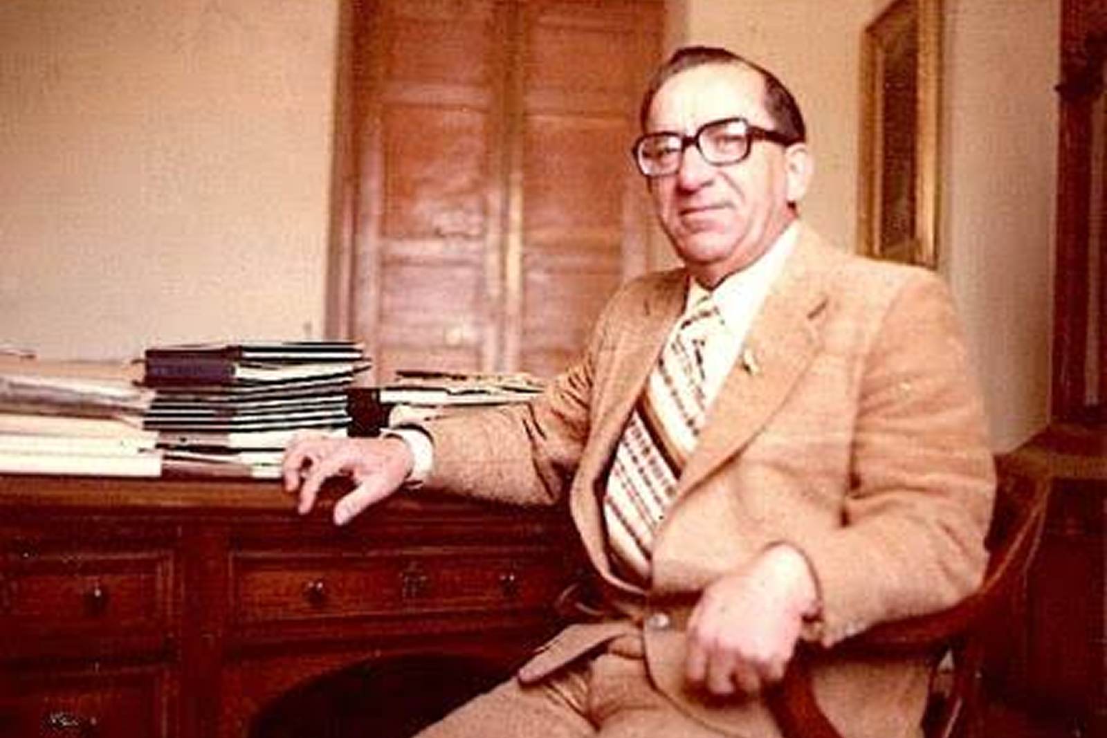 Colour portrait of a Maltese politician of the integration era, in a light tan suit and thick-rimmed glasses, seated beside a wooden desk stacked with books and papers, panelled shutters behind him.