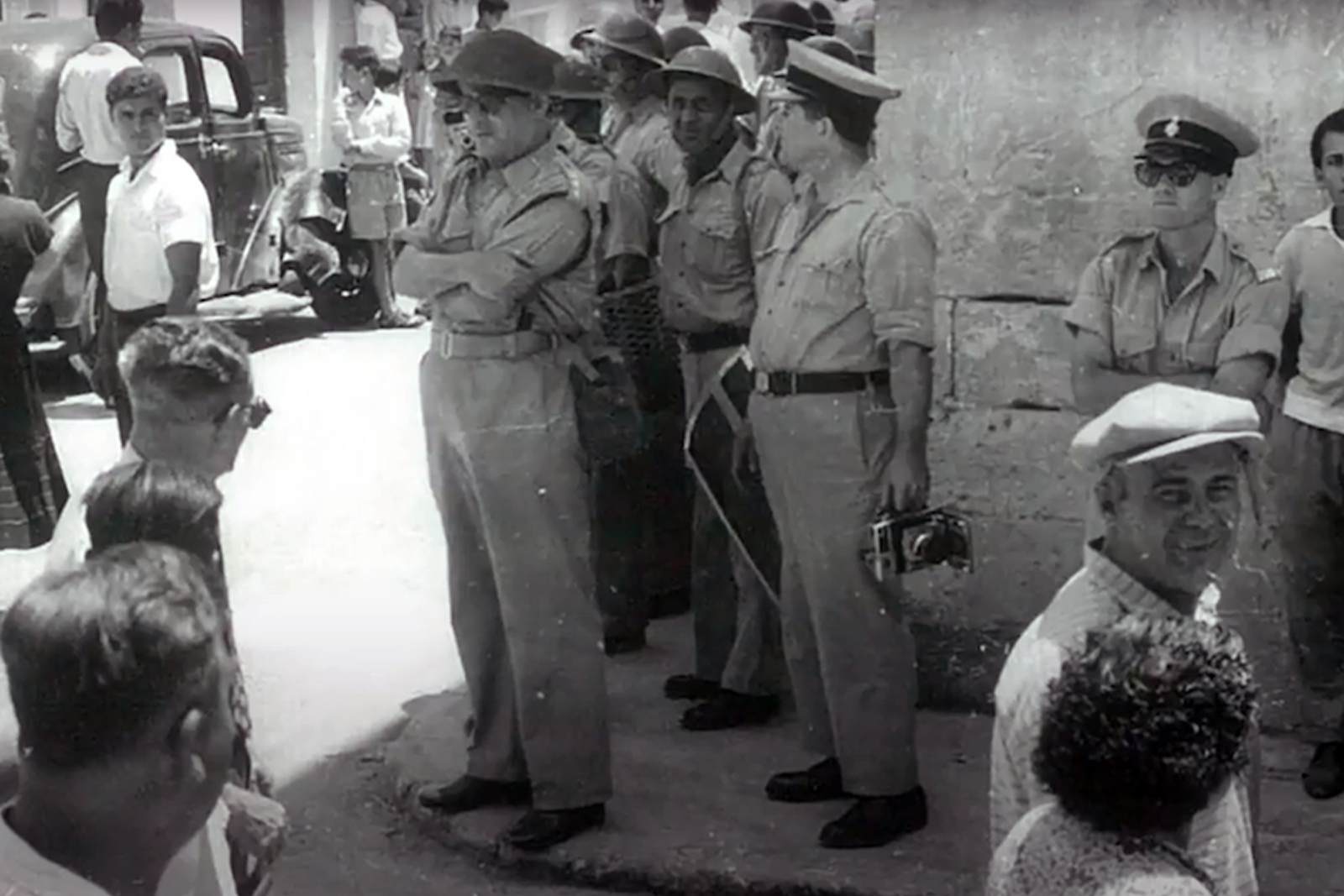 Black and white street scene from 1950s Malta: British servicemen in khaki uniforms and helmets standing in a narrow village street, one holding a press camera, with a police officer and local onlookers gathered beside a parked vintage car.