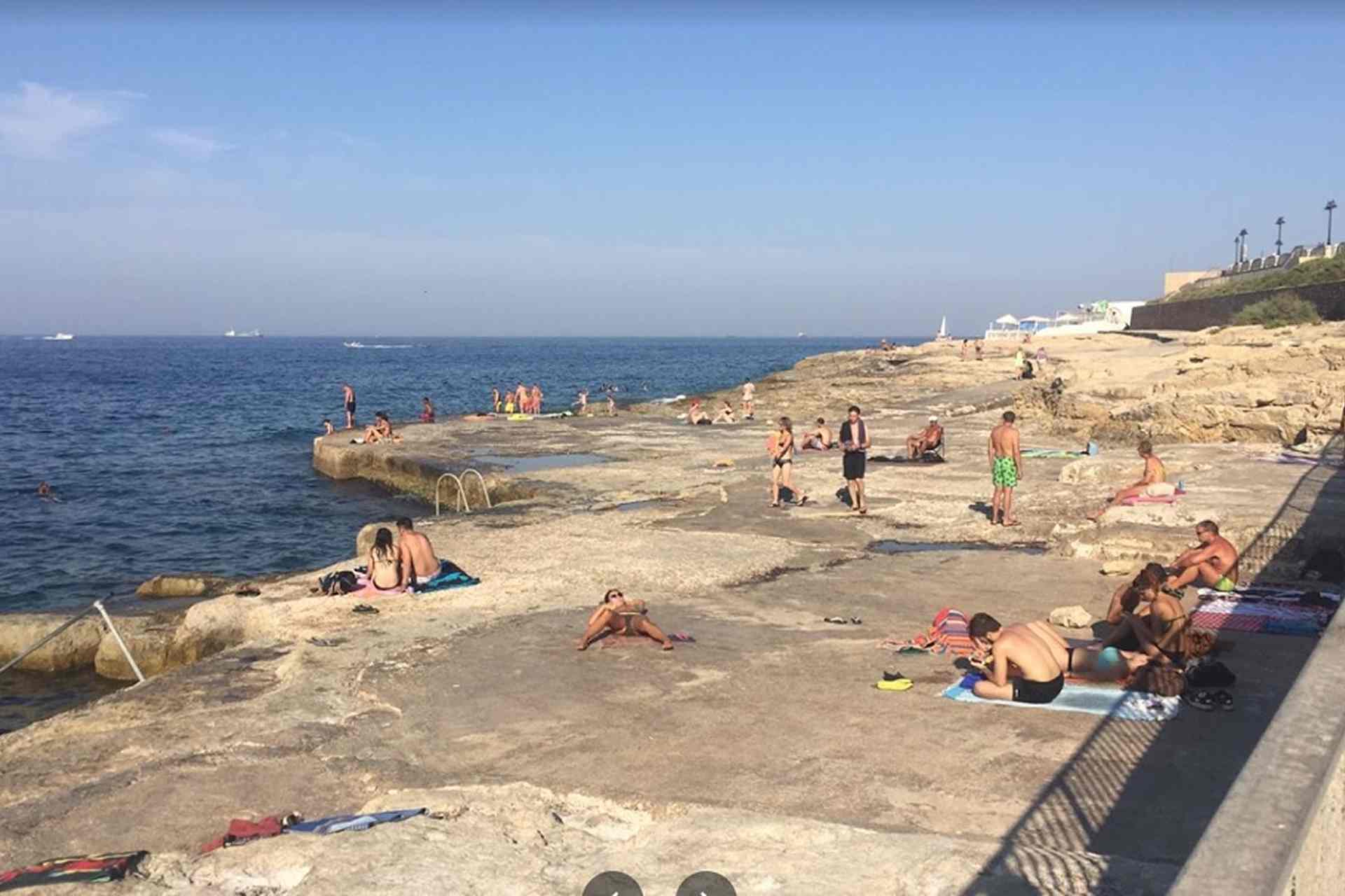 Rocky swimming area at Qui-Si-Sana, Sliema, Malta, with sunbathers on the flat rocks and people enjoying the sea on a sunny day