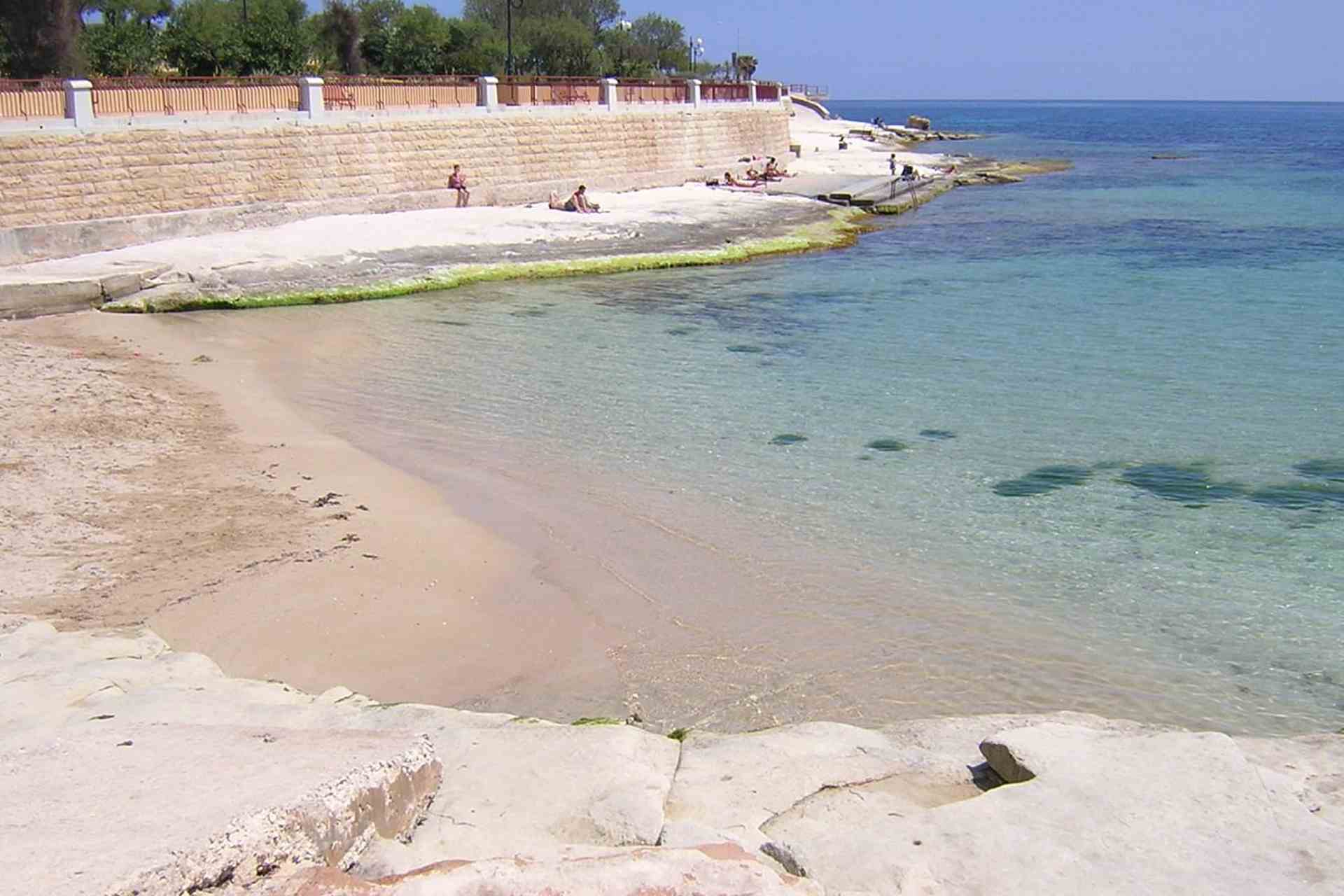 Clear beach with gentle waves and shallow water at St Thomas Bay, Malta, bordered by sandy shore and stone wall. A few people are relaxing by the water