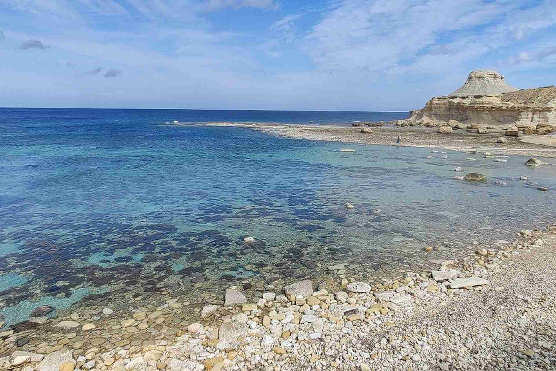 Pebble beach at Xwejni Bay, Gozo, Malta, with crystal clear water, rocky coastline, and dramatic limestone formations under a partly cloudy sky