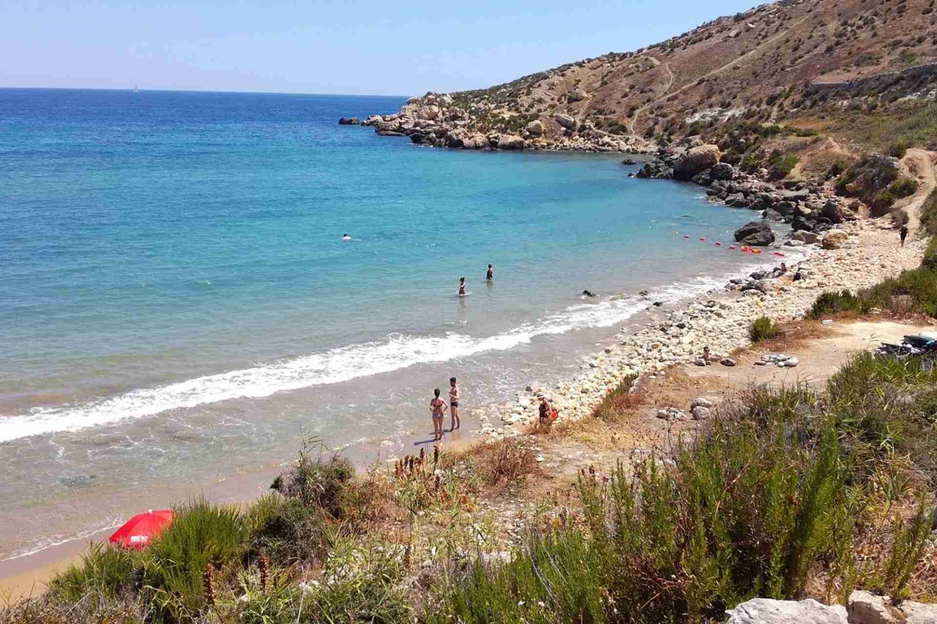 Remote sandy beach at Imgiebah Bay, Malta, with turquoise water and rocky hills in the background. A few people are swimming and relaxing on the shore.